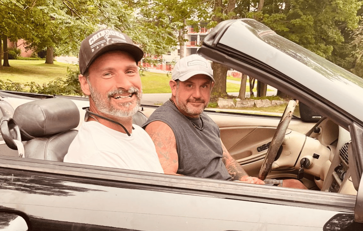 Tarzan Kay's boyfriend and brother smiling together in a black convertible car on a sunny summer day