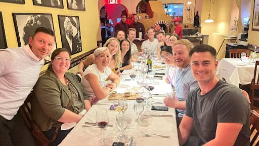 A group of people are seated around a long, white-tableclothed table in a restaurant. There are wine glasses, plates, and cutlery on the table. The group is smiling and looking toward the camera. The restaurant has yellow walls, framed photos, and warm li