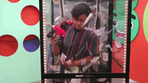 A GIF of a guy wearing a red and green striped shirt inside a money blowing machine trying to grab the cash while holding a red robot in his hand.