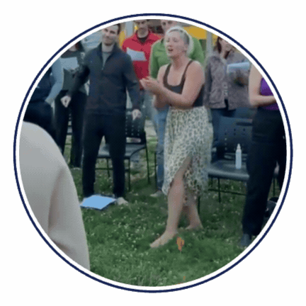 Animated GIF of Tarzan singing barefoot on grass, wearing a black tank top and an animal print skirt, with a group of people standing in a circle with her, singing along.
