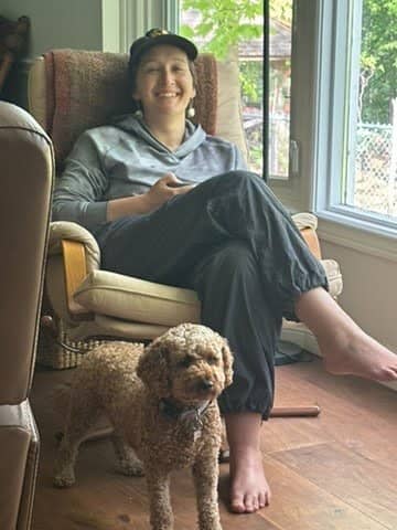 Tarzan Kay relaxing barefoot in a cozy chair by a sunlit window with a small curly-haired dog standing nearby, capturing a warm and peaceful work-from-home or slow living moment.