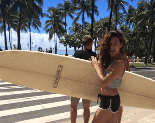 Woman carrying a surfboard across a sunny crosswalk lined with palm trees, smiling at the camera on a tropical beach street, capturing a carefree summer surfing lifestyle.