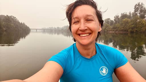 Tarzan Kay smiling and taking a selfie while paddling on a calm river surrounded by trees on a misty day