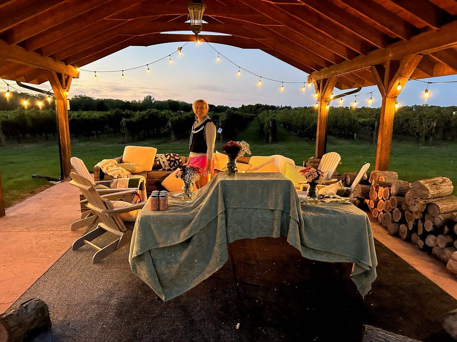 Cozy outdoor pavilion scene at dusk with warm string lights hanging from a wooden roof structure. Tarzan stands near the center, surrounded by cushioned chairs, a table covered with a green tablecloth, and rustic details like flower arrangements and stack