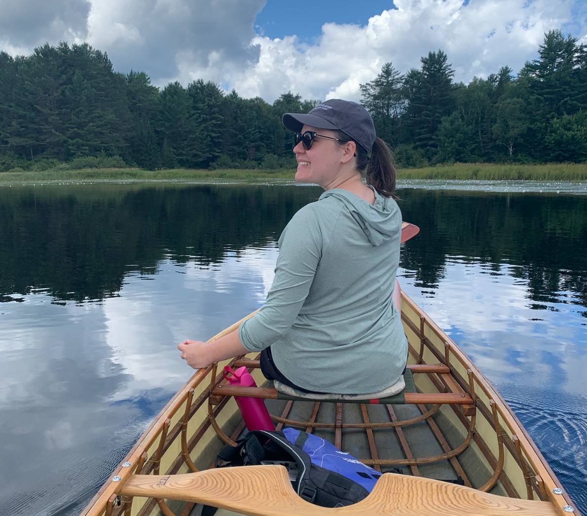 Woman smiling while canoeing on a peaceful lake surrounded by a lush green forest under a partly cloudy summer sky.