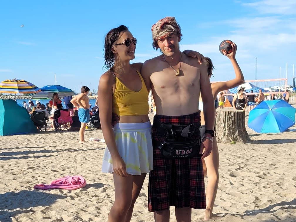 Tarzan wearing a yellow top and white skirt stands on a sandy beach with their arm around Quentin wearing a plaid kilt, a head wrap, and a cross-body pouch. They are both smiling. Behind them are beach umbrellas, people sitting in chairs, and others walki