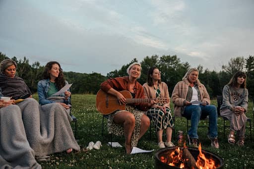 A group of six people sit outdoors in a grassy field around a campfire. Tarzan is in the middle is playing an acoustic guitar while others sit beside them, some holding papers or covered with blankets. The atmosphere appears relaxed and communal, with tre