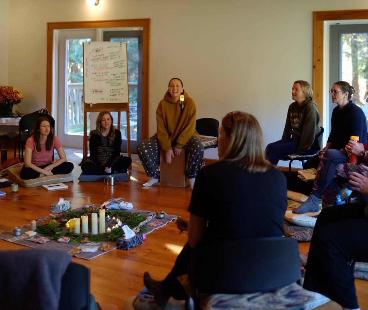 A group of women sit in a circle on cushions and chairs in a warm, sunlit wooden room, singing In the center, a centerpiece of evergreen boughs, lit candles, and small objects is laid out on a woven rug. Tarzan is smiling, in a mustard hoodie, and sits on