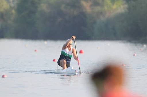 Tarzan Kay paddling intensely on a calm river during a canoe or kayak race with buoy markers visible on a sunny morning