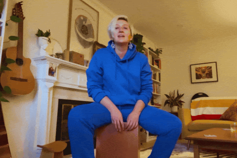 Tarzan in a bright blue sweatsuit energetically speaking while sitting on a stool singing in a cozy, stylish living room with guitars, bookshelves, and colorful decor in the background.