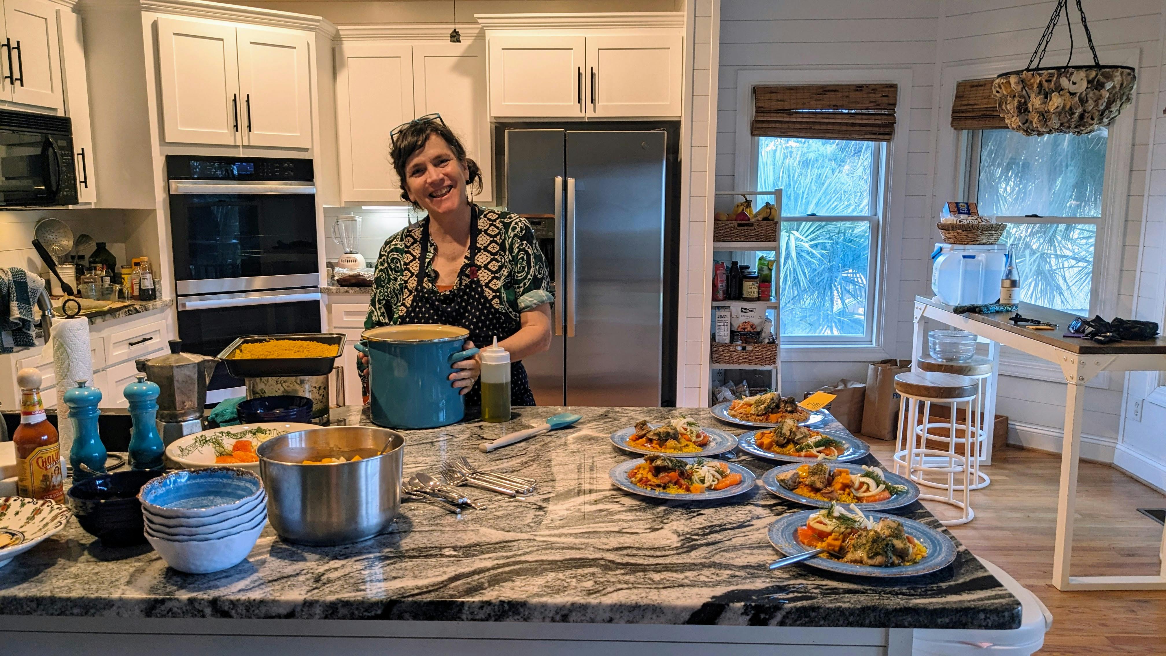 a smiling brunette woman in a kitchen, in front of several plates of food she's prepared