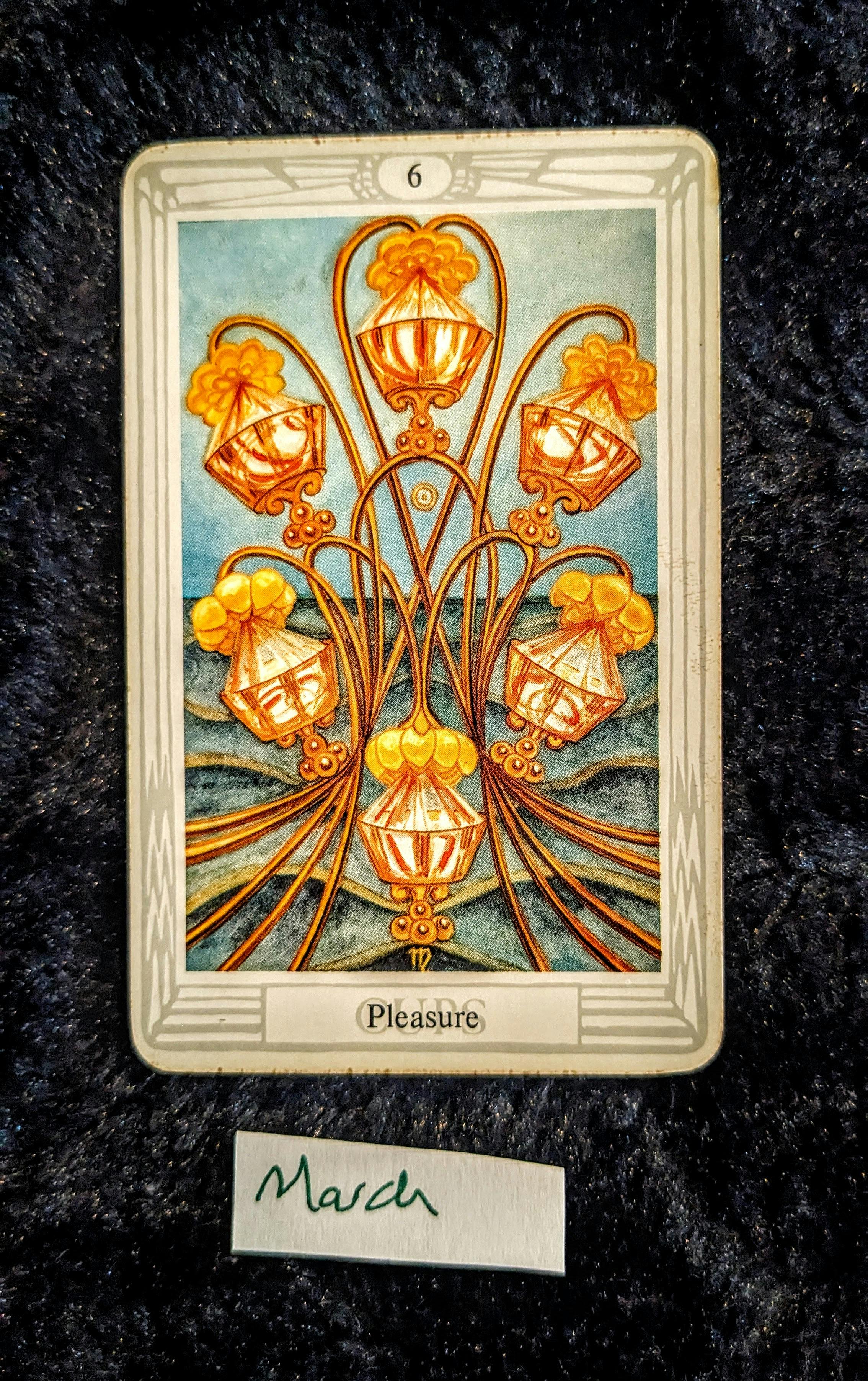 a photo of the 6 of cups tarot card from the thoth deck on a black background.  on a slip of paper under the card, the word March is hardwritten in ink.  the card depicts six golden cups joined in harmony against an ocean-like background.  on the bottom of the card reads the word "Pleasure"