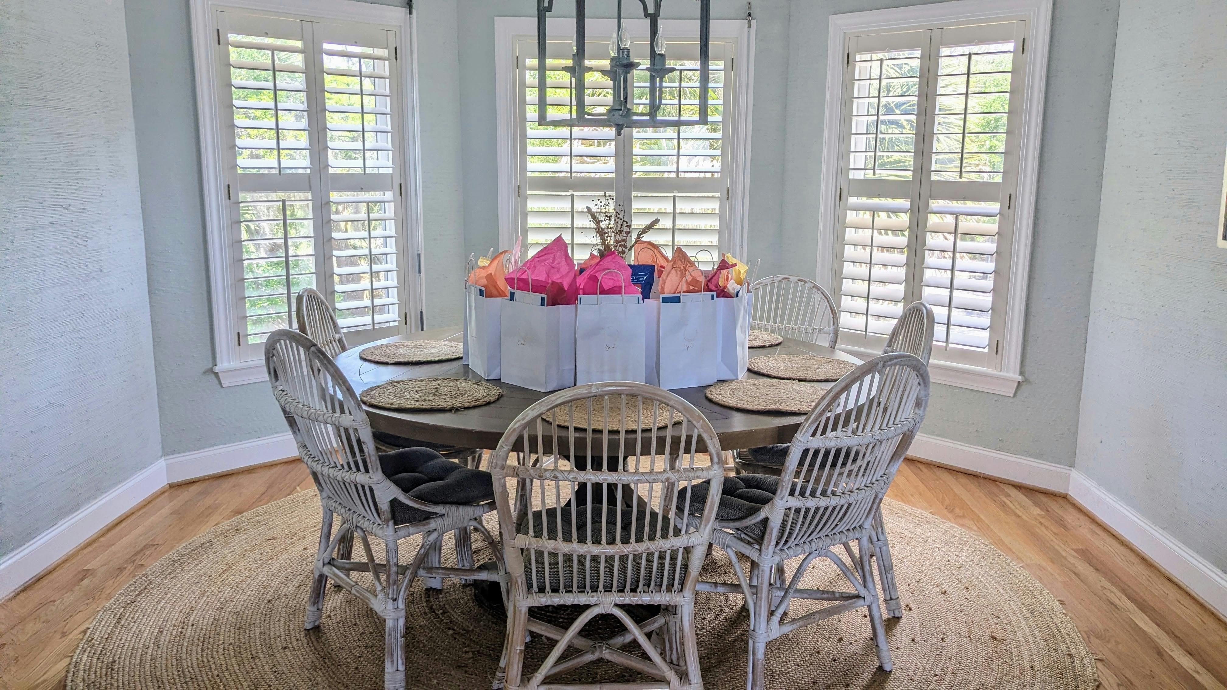 a photo of a table in a beach house surrounded by chairs, with a circle of gift bags on it