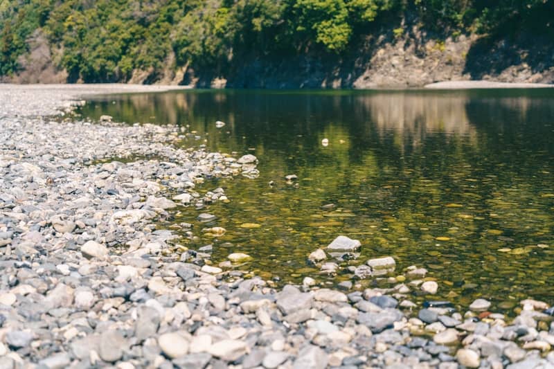 Rocky riverbank beside calm water with green trees and cliff.