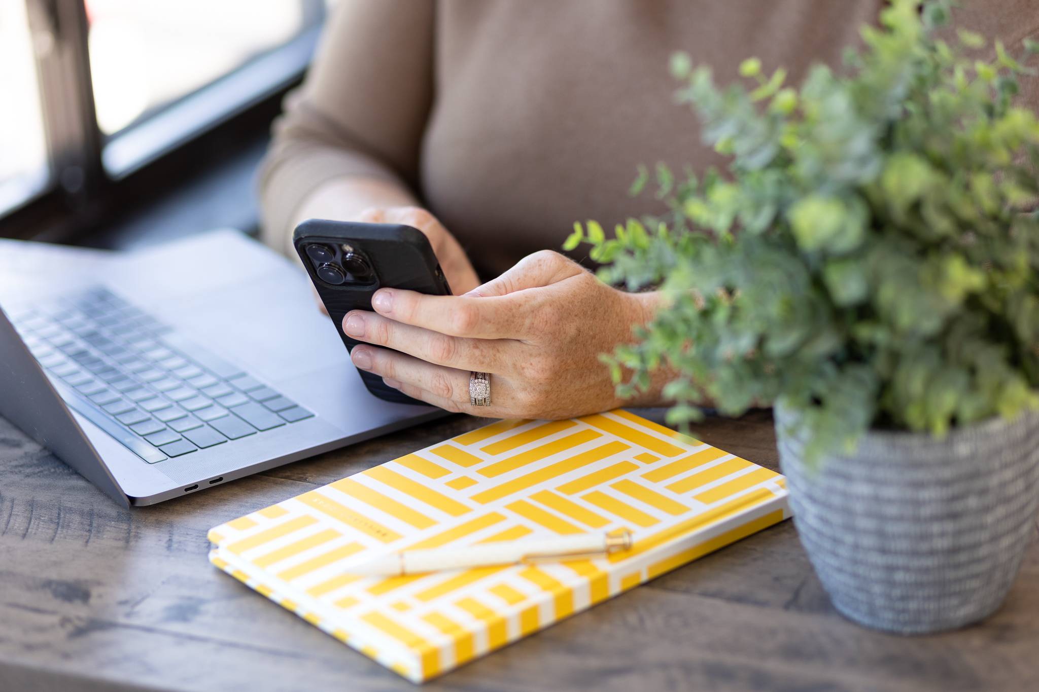 Hand holding a phone next to a laptop and yellow patterned notebook