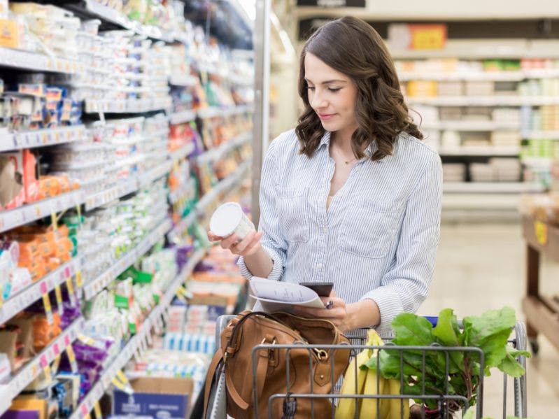 Woman grocery shopping in store with list and grocery cart filled with produce.