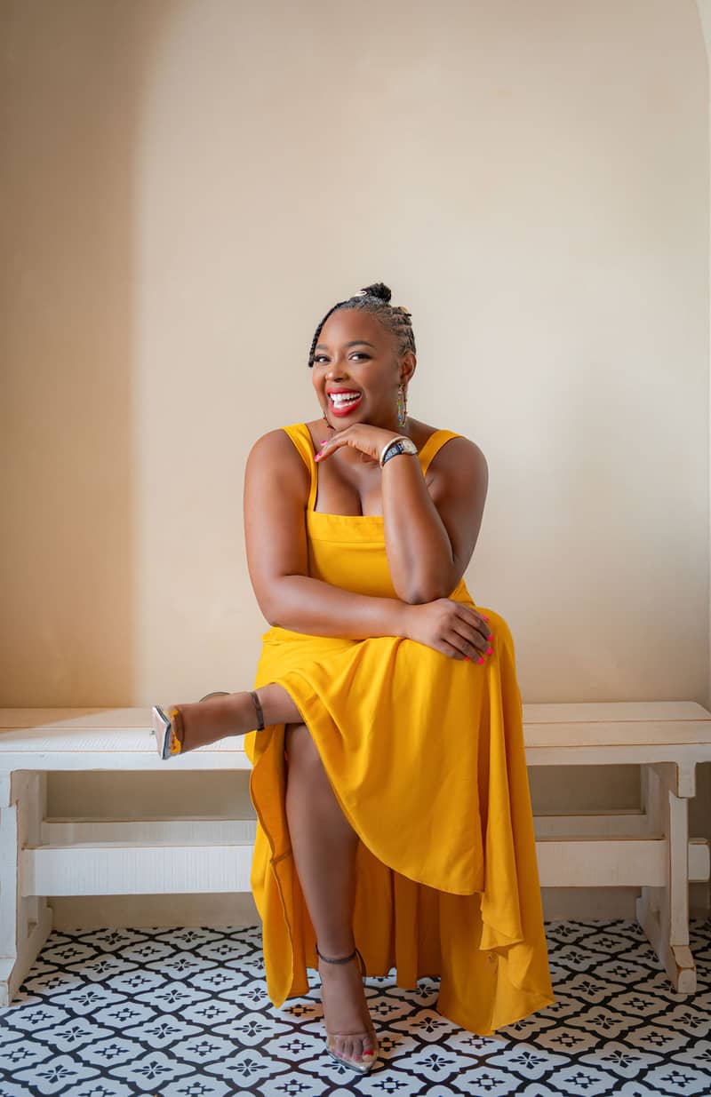 A person with braided hair is sitting on a bench against a beige wall. They are wearing a bright yellow dress and transparent high heels. The person is smiling and resting their hand on their chin, displaying colorful earrings and a watch. The floor has a black and white geometric pattern.