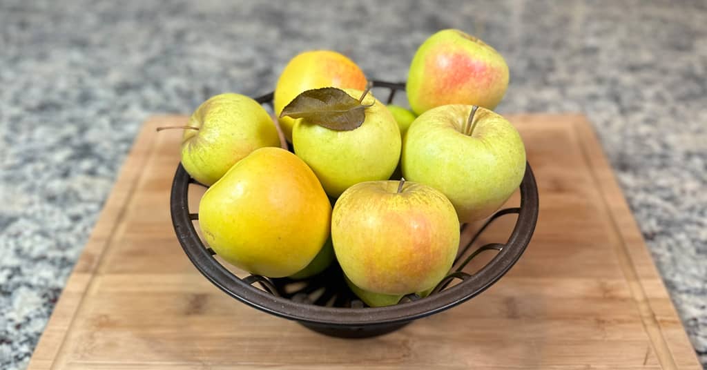 Yellow and red apples sitting in a metal bowl on a cutting board on a counter.