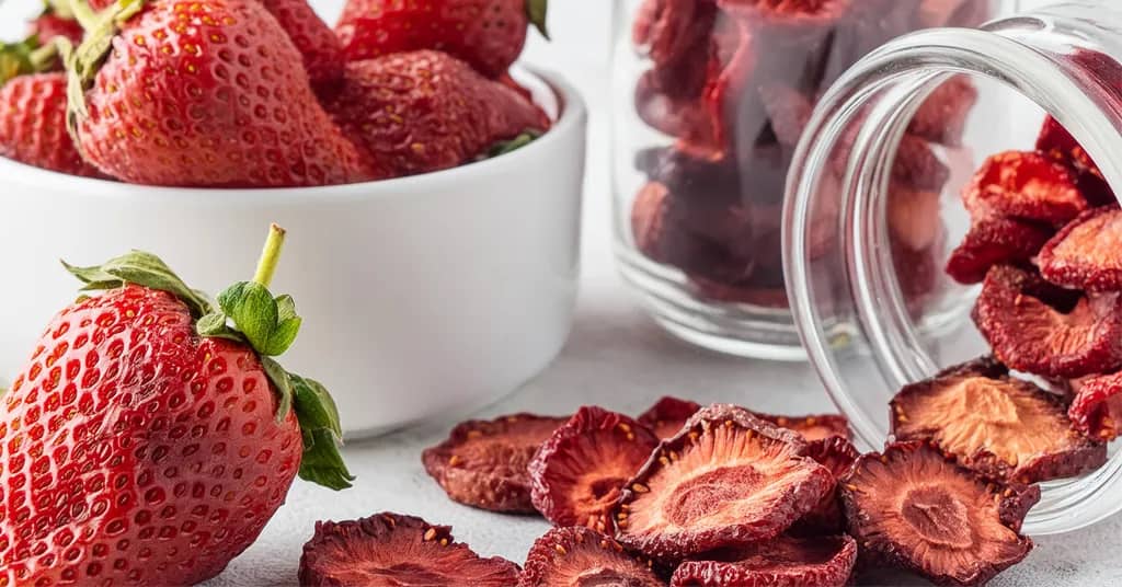 Dehydrated strawberry slices spilling out of a mason jar with a bowl full of fresh strawberries.