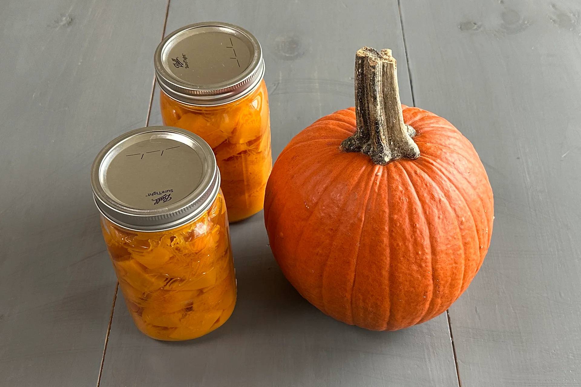 Two mason jars of pumpkin cubes and a sugar pie pumpkin sitting on a gray surface.