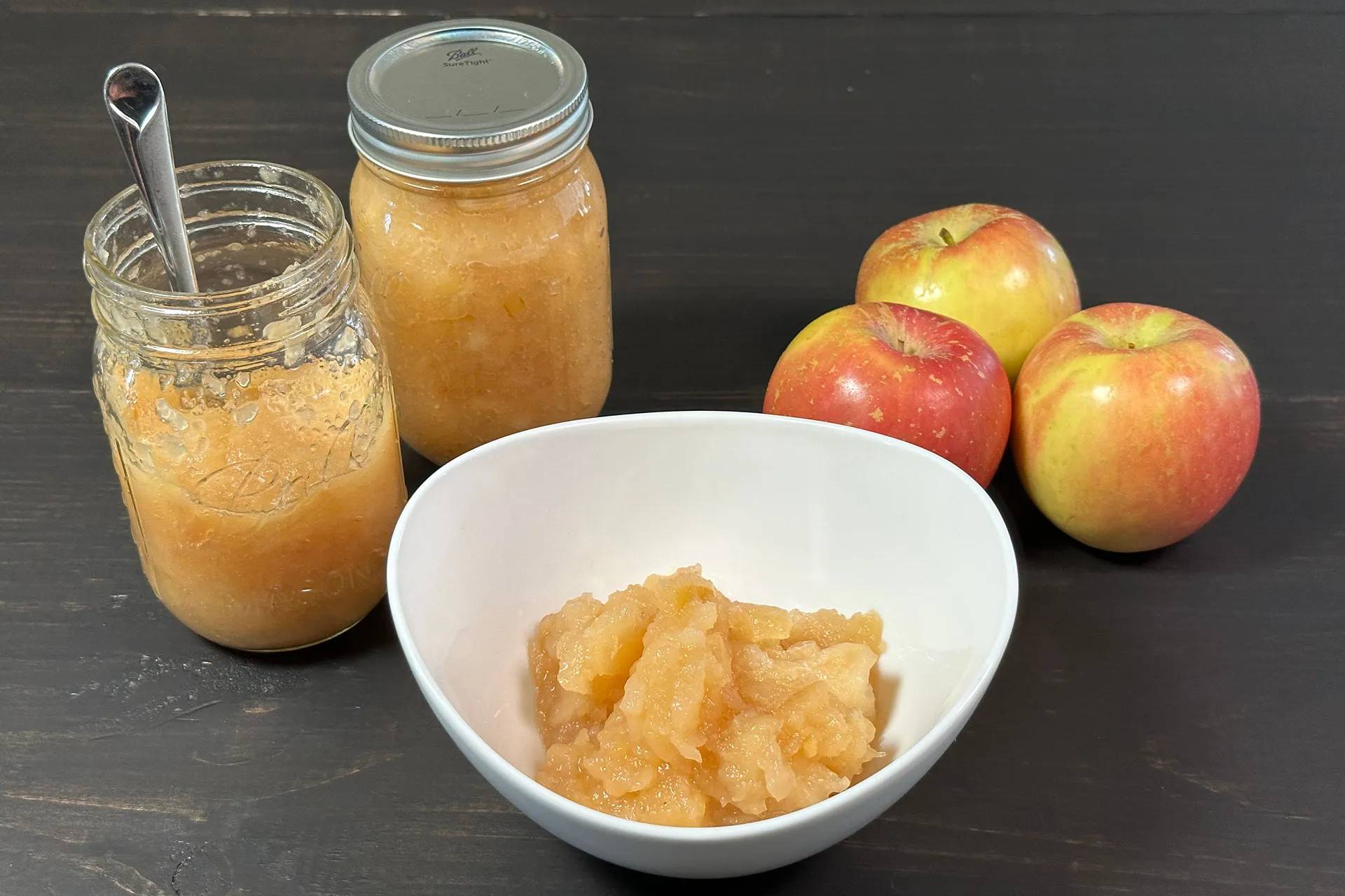 Apple sauce in a white bowl and in two mason jars with apples on the side.