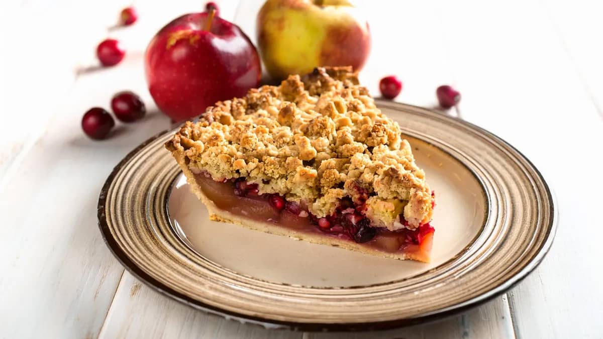 Apple pear cranberry pie slice on a plate with fresh apples and cranberries in the background.