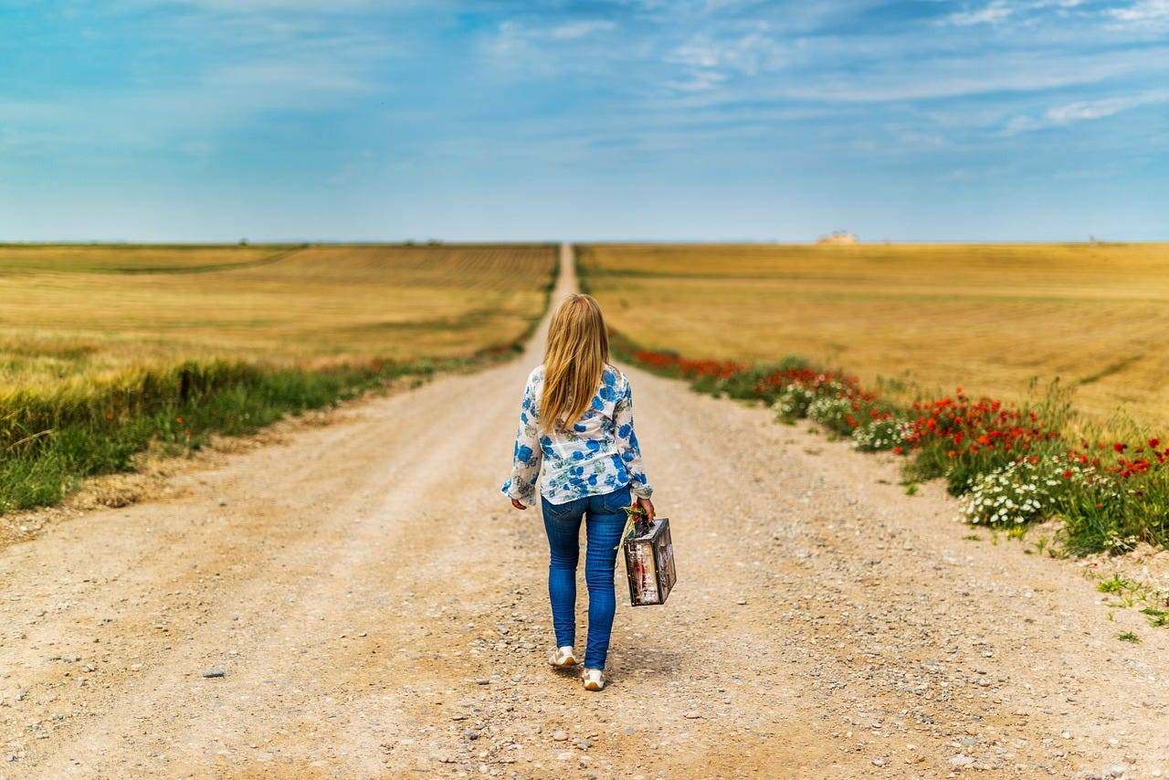 A woman walking down a dirt road with a suitcase in hand.