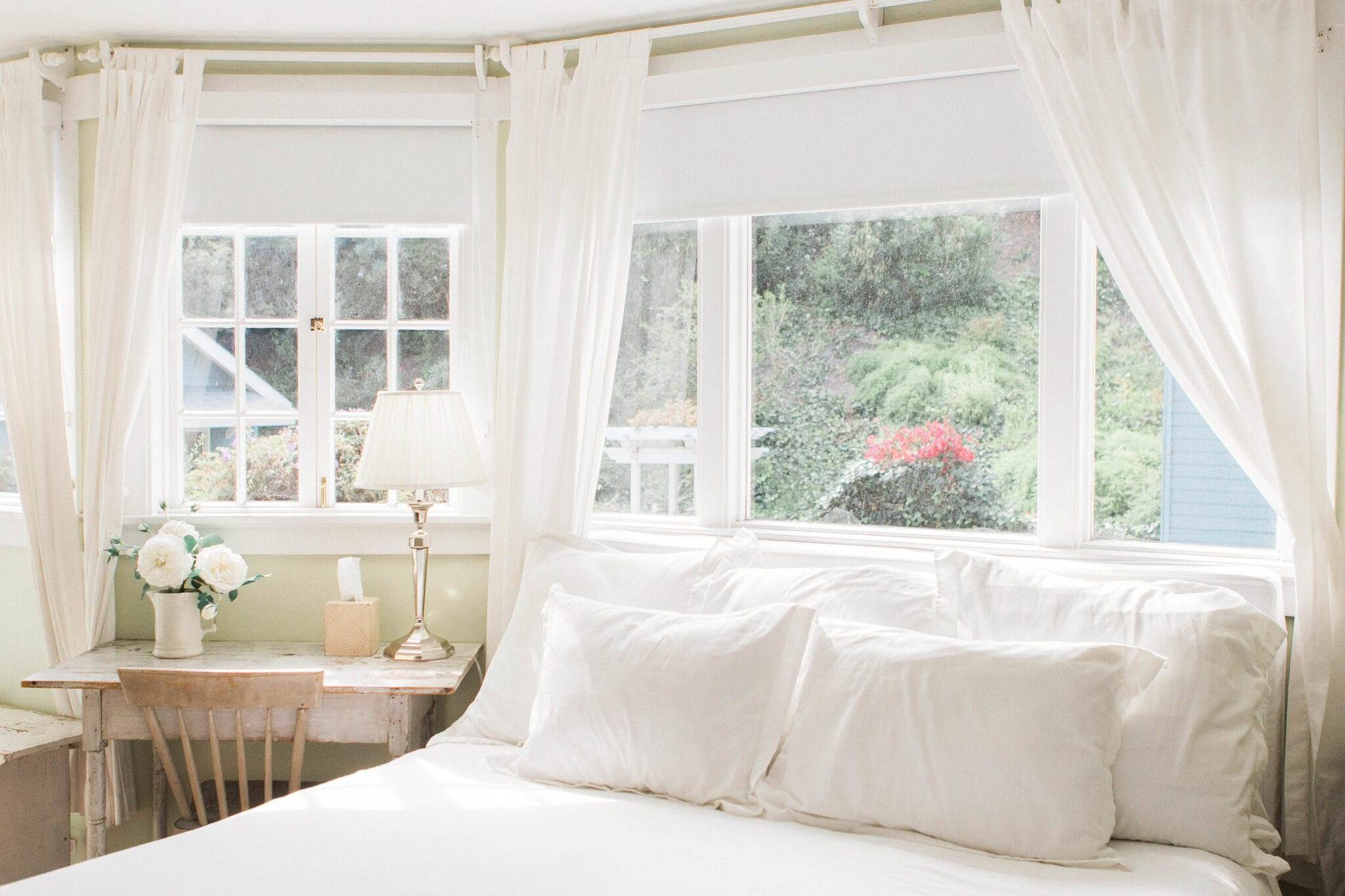 A bright bedroom looking at the bed, with windows behind it and a desk to the left. The windows are draped with long white curtains. The desk has flowers in a vase, a tissue box, and a lamp on it, as well as a chair pushed underneath.