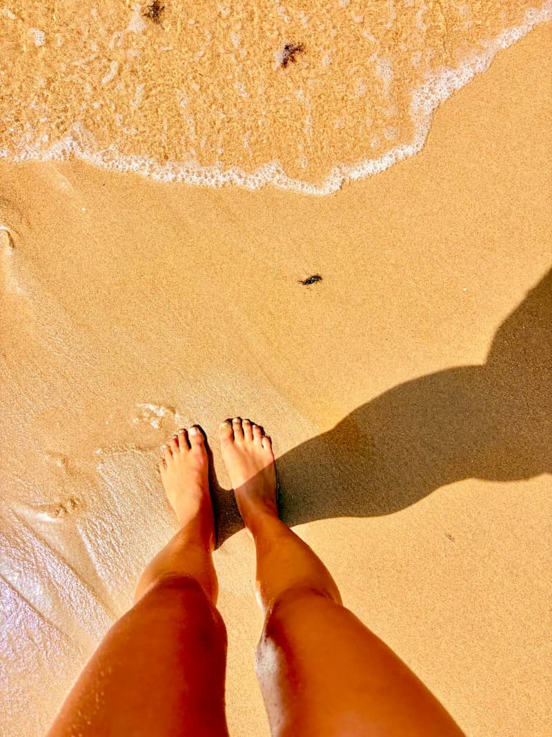 A person standing on wet golden sand with a long shadow cast by the sun