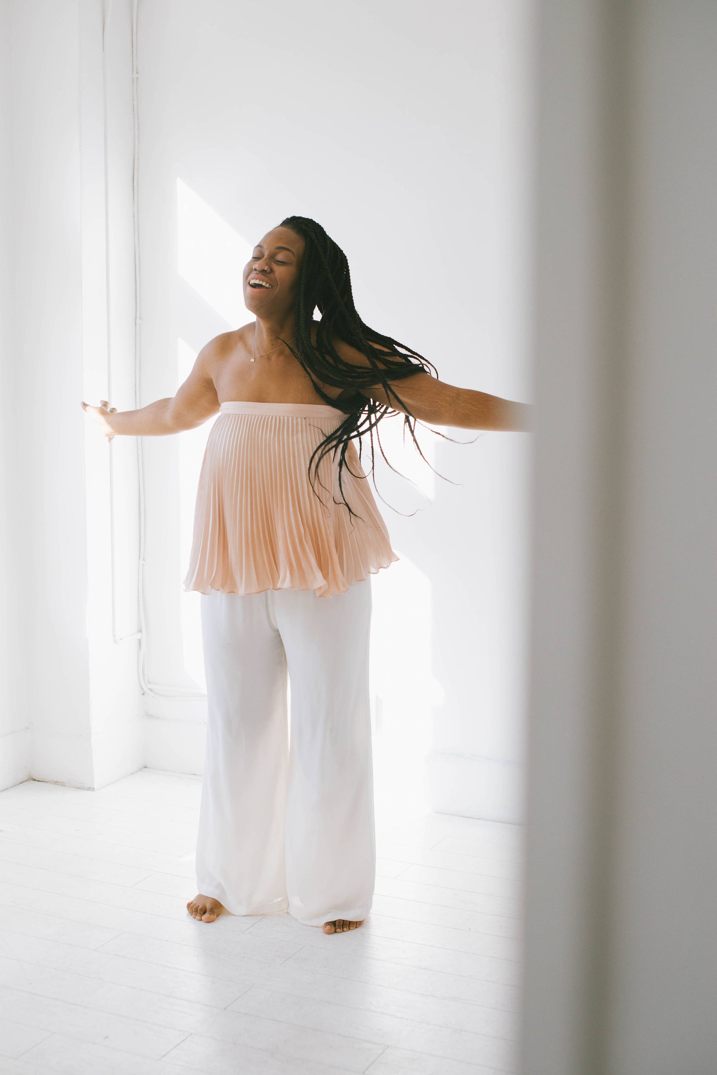 Black woman in a pink strapless shirt and white, wide leg pants with arms outstretched and a soft smile on her face