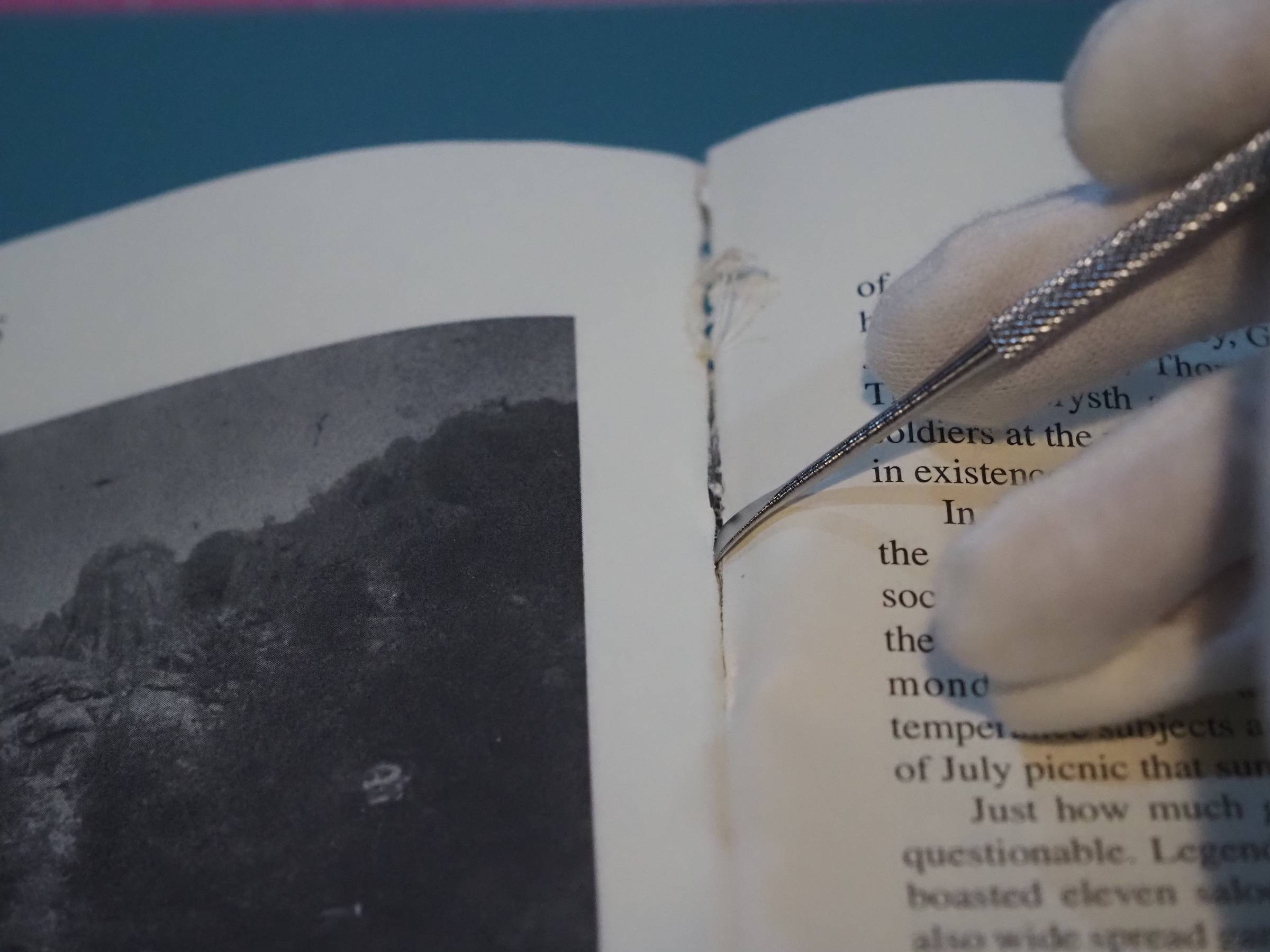 a white gloved hand holds a jewelers scraping tool to pull apart the glue and binding on a book.
