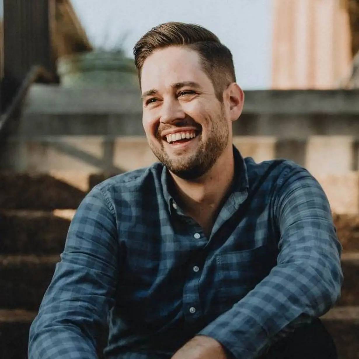 A man sitting on outdoor steps, smiling broadly. He has short hair and a beard, and he is wearing a blue plaid shirt. The background is softly blurred, suggesting a sunny day.