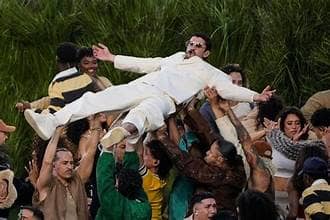 Bad Bunny after a trust fall into a crowd of backup dancers and performers at Super Bowl 60. He is wearing an ivory suit and dark shades while the crowd is dressed in bright, modern clothing.