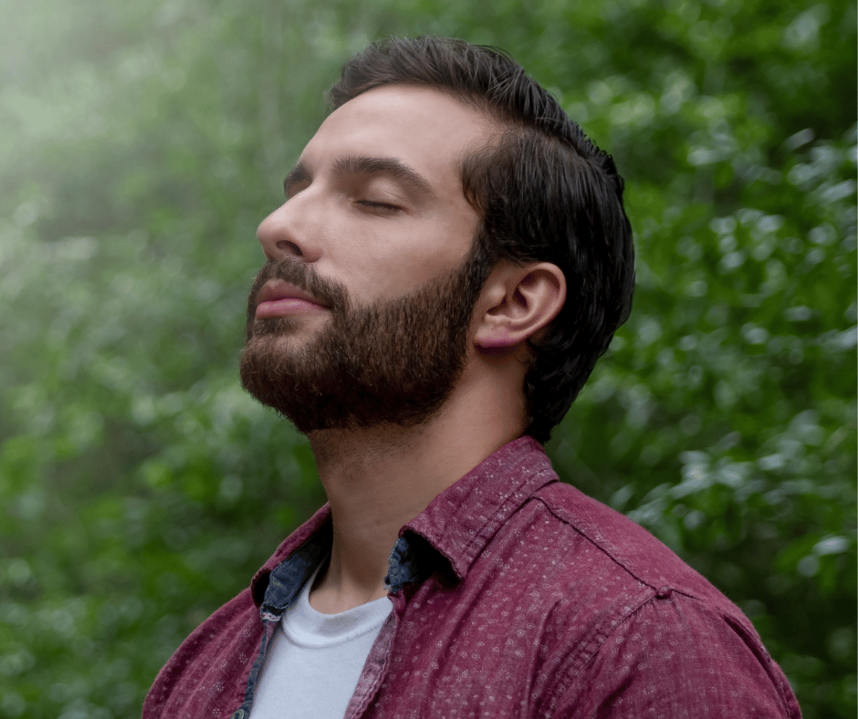 Calm dark-haired man with beard with closed eyes, against background of greeneryc