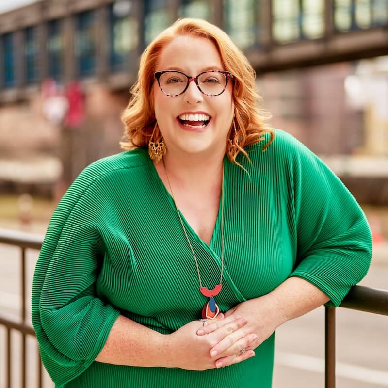 A woman with red hair is smiling and leaning on a railing. She is wearing glasses, a green dress, and colorful jewelry. The background is blurred, showing an outdoor scene with a building.