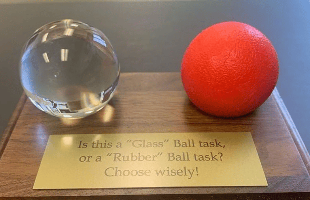a desktop plaque with the words "is this a glass ball task or a rubber ball task? Choose wisely! and a glass ball and an orange rubber ball.
