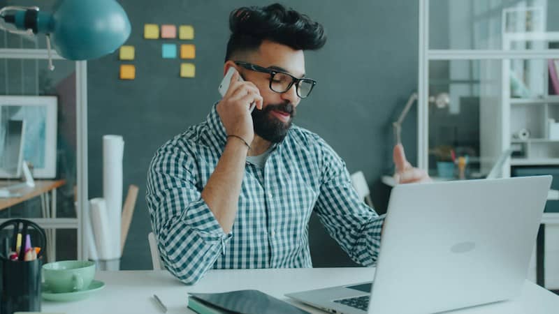 A man with a beard and glasses talking on a phone at a desk