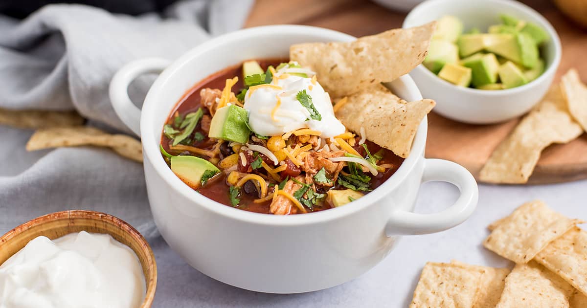 close-up of a white bowl full of enchilada soup, loaded up with diced avocados, shredded cheese, corn, cilantro, sour cream, and garnished with tortilla chips 