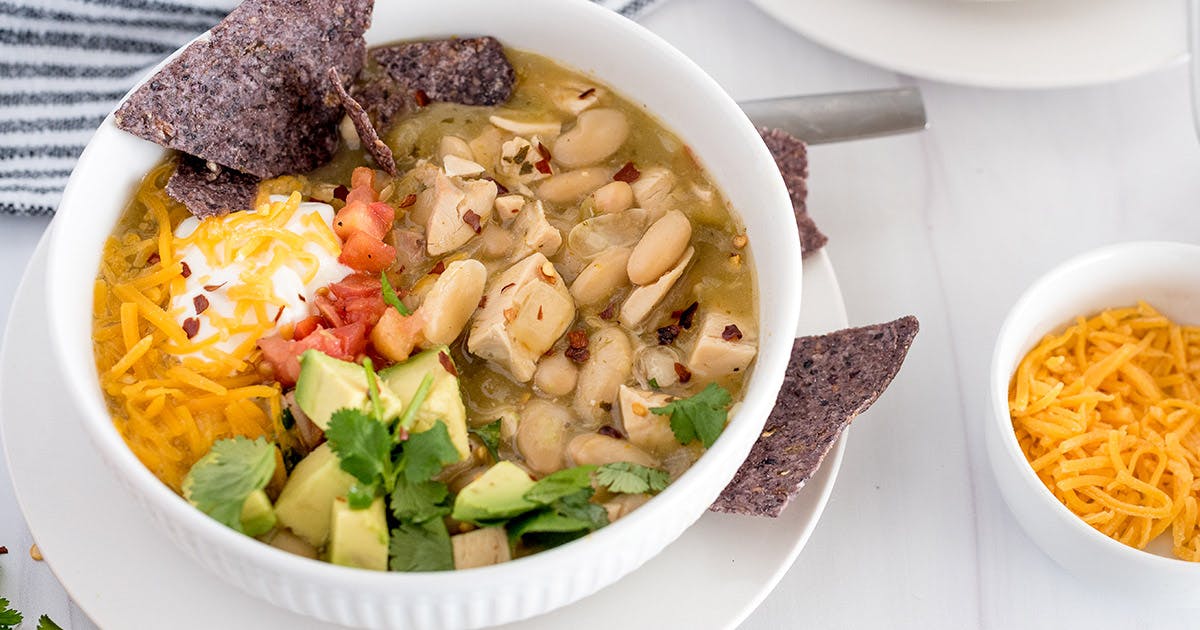 close-up of a white bowl filled with INstant Pot White Chicken Chili, garnished with blue corn chips, shredded cheese, and green avocado