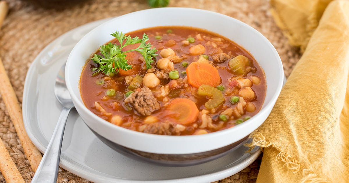 close-up of a white bowl filled with Vegetable Beef Soup, studded with carrots, celery, peas, potatoes, and rice