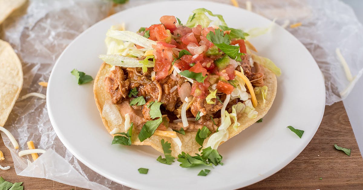 45 degree close up of a tostada, all plated up on a white plate on a yellow corn tortilla, topped with sweet pork, tomatoes, and cilantro.