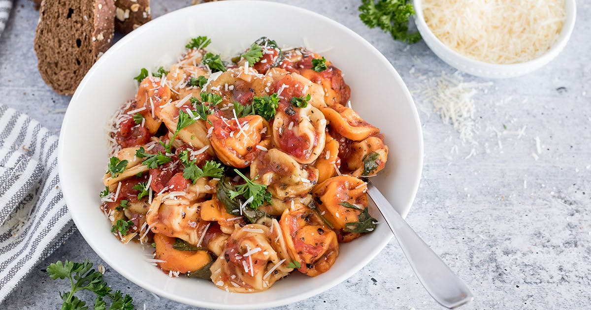 Close up picture of Instant Pot tortellini topped with fresh parmesan and parsley, placed in a white bowl. Additional parmesan and slices of wheat bread in the background.