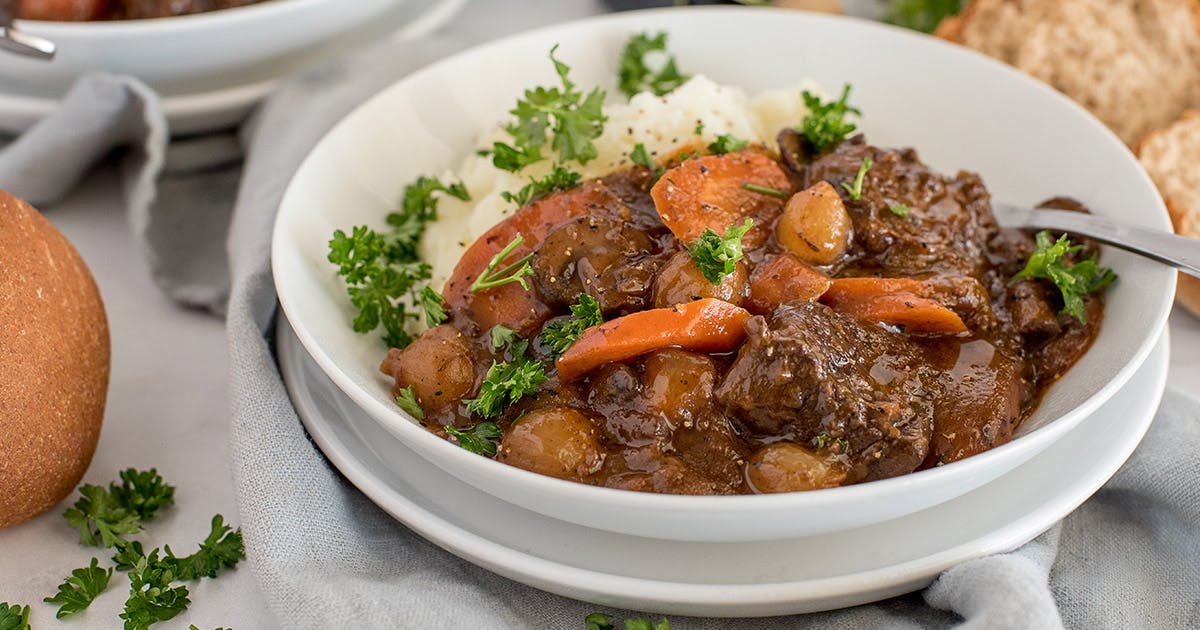  a white bowl of Beef Bourguignon with mashed potatoes, and a roll in the background