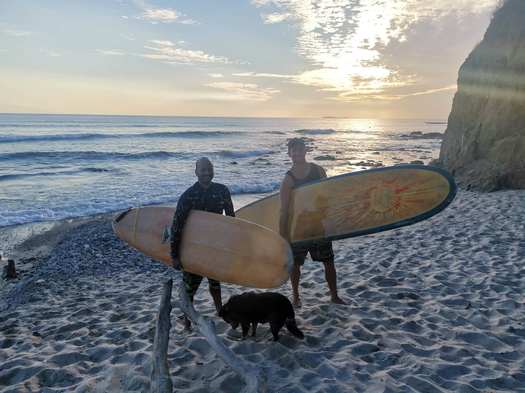 Allan and Workaway host Juan standing on the beach in front of the ocean at sunset with two big yellow surf boards under their arm and a black dog next to their feet.