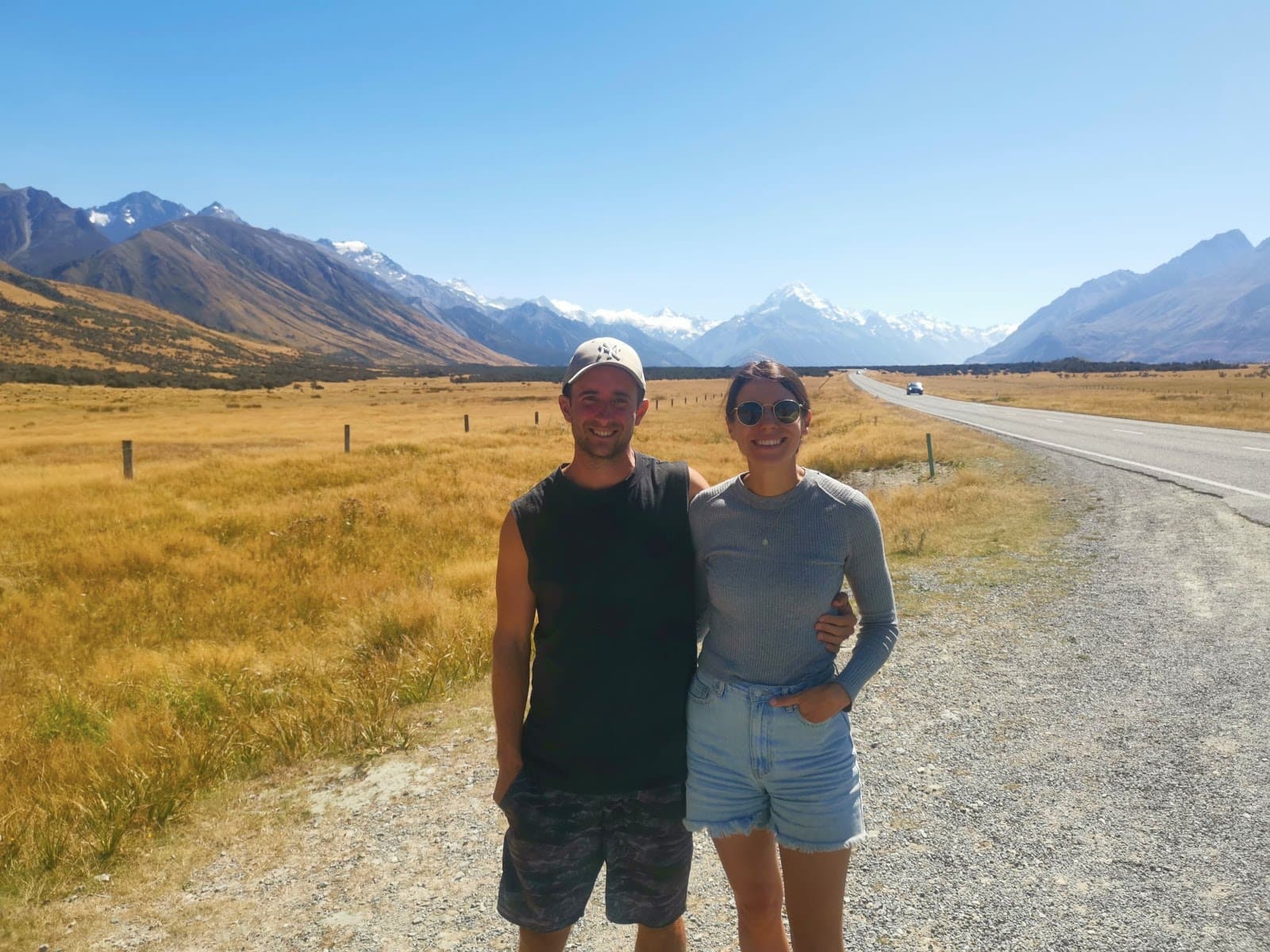  Allan and Katharina pose for a photo on the side of a road in New Zealand's Mackenzie Basin, with the snow-capped peak of Mount Cook and the Southern Alps visible in the background.