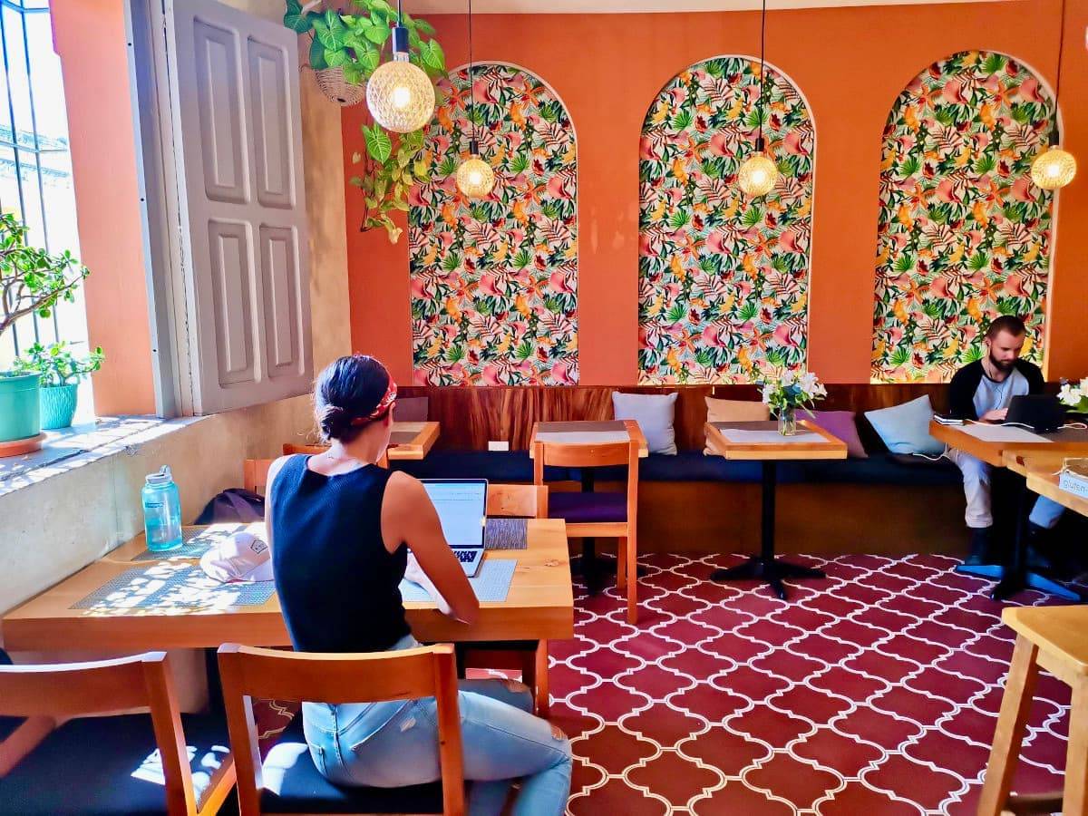 Katharina sits at a wooden table in a brightly lit cafe with tropical decor, working on her laptop during a slow travel trip.