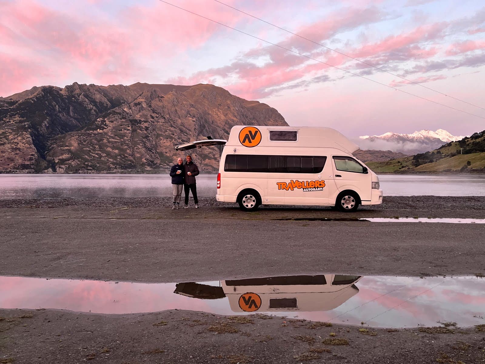 Katharina and Allan stand together holding mugs next to their white Travellers Autobarn campervan on a gravel shore of Kidds Bush campsite near Wanaka, with their reflections mirrored in a calm puddle foreground and a stunning pink sunset sky over mountai