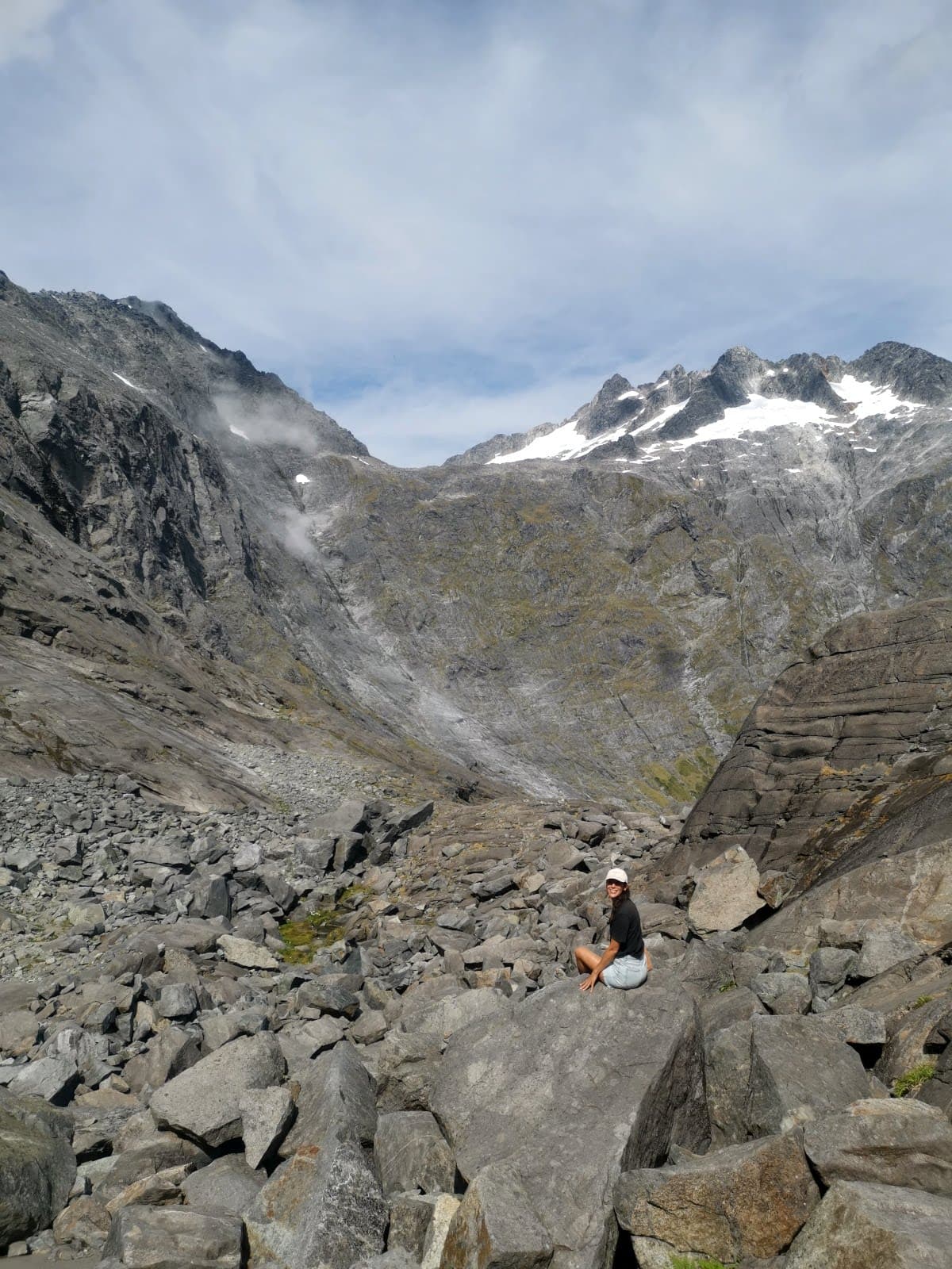 Katharina sitting on a grey rock surrounded by rock and a snow cap mountain peak on the Gertrude Saddle hike near Milford Sound.