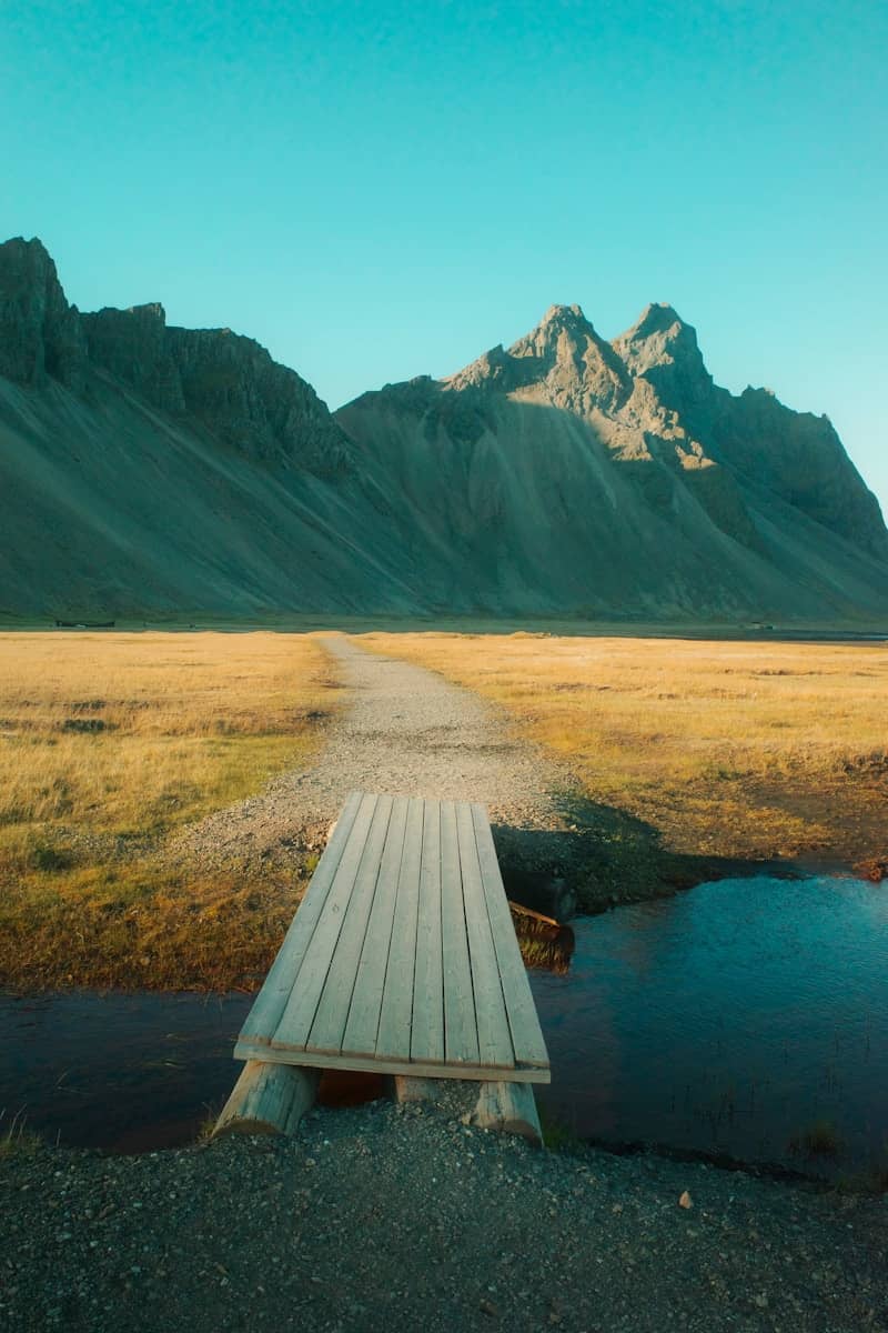 Wooden bridge over stream with golden fields and rugged mountains.