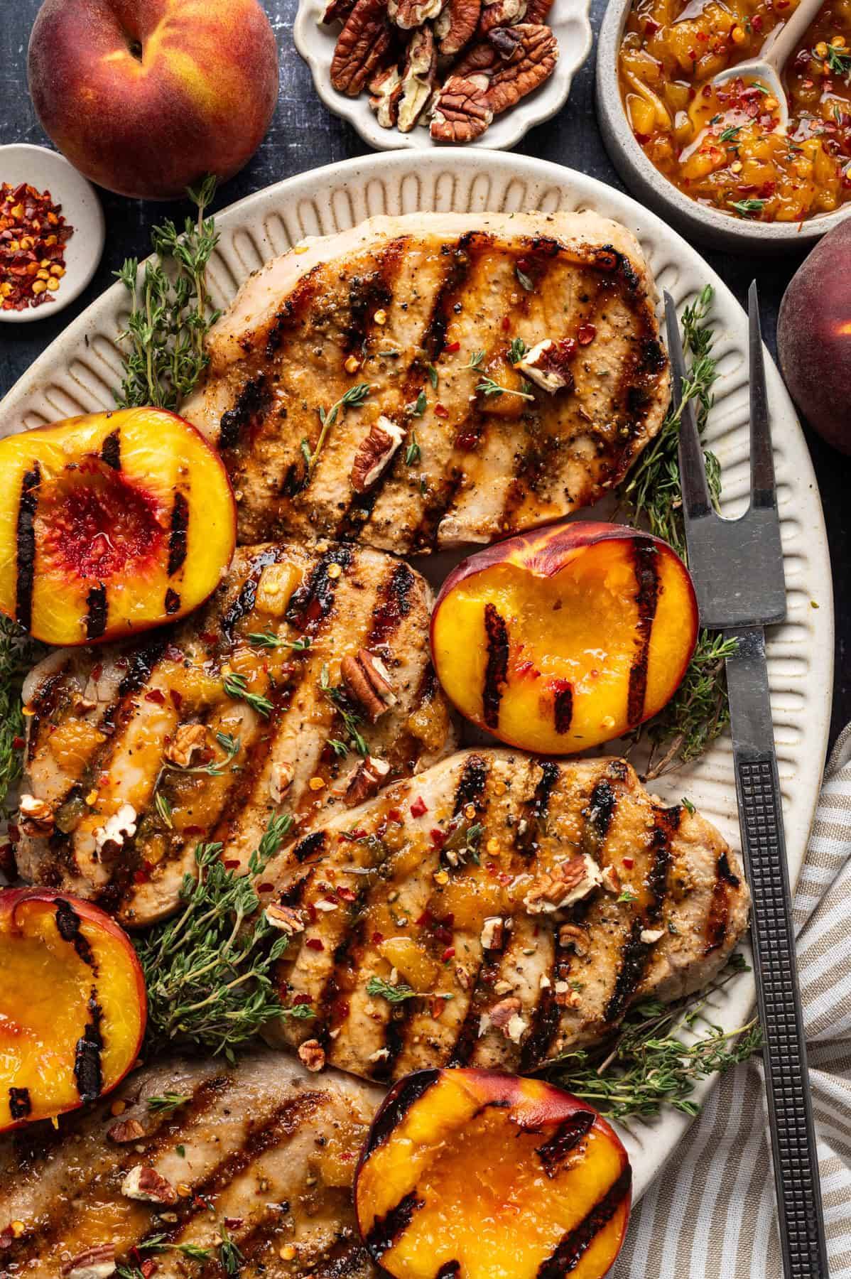 Bourbon Glazed Grilled Pork Chops with Grilled Peaches, Peach Sauce, and Pecans on a white platter.